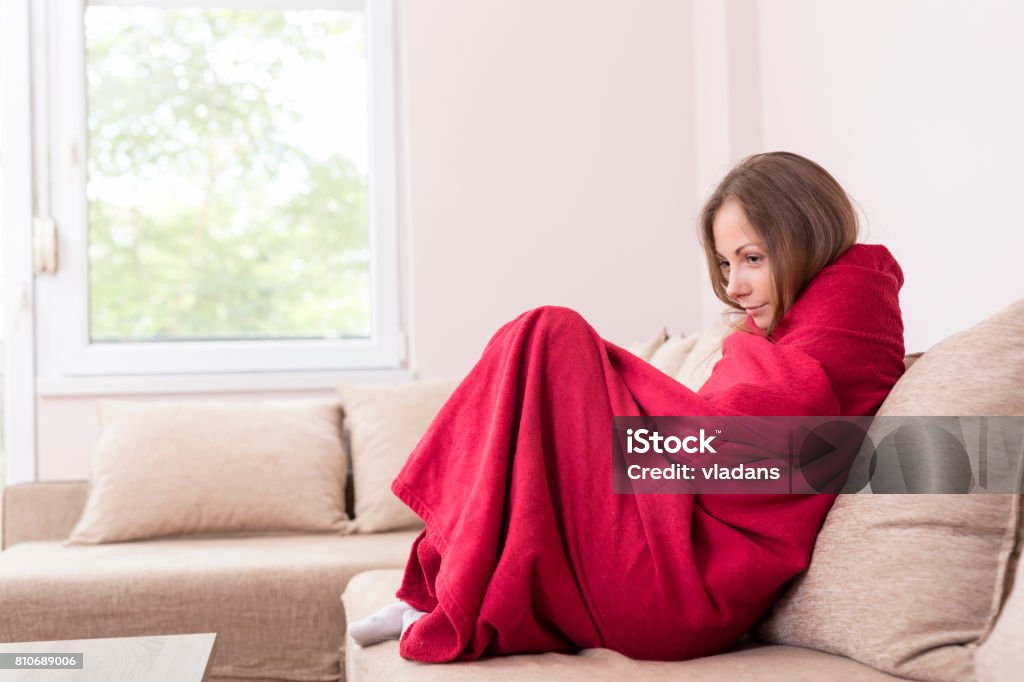 Cold symptoms Young woman sitting on a couch covered with blanket, having a fever. Cold Temperature Stock Photo