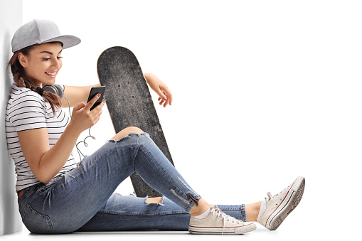 Teenage girl with a skateboard looking at a phone and leaning against a wall isolated on white background