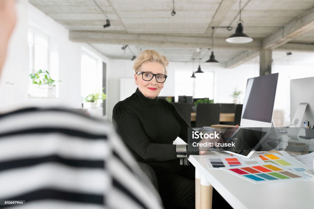 Senior businesswoman using a digital tablet in the office Senior interior designer sitting at the desk in the office, pointing at digital tablet and talking with her coworker. 60-69 Years Stock Photo