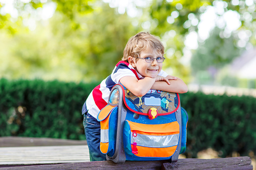 Happy little kid boy with glasses and backpack or satchel on his first day to school . Child outdoors on warm sunny day, Back to school concept. Kid with traditional schoolbag in German Schultuete