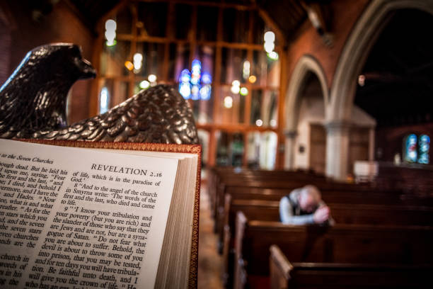 Man praying on pew inside English Anglican church with focus on bible in foreground Close up image of the Holy Bible open at the book of Revelation on the altar of an Anglican church. The pews and stained glass windows of the interior of the church are blurred out of focus in the background, and warm orange sunlight is streaming in through the windows. On one of the front pews a young man is bowed in prayer, and he too is blurred out of focus. Horizontal colour image with copy space. anglican eucharist stock pictures, royalty-free photos & images