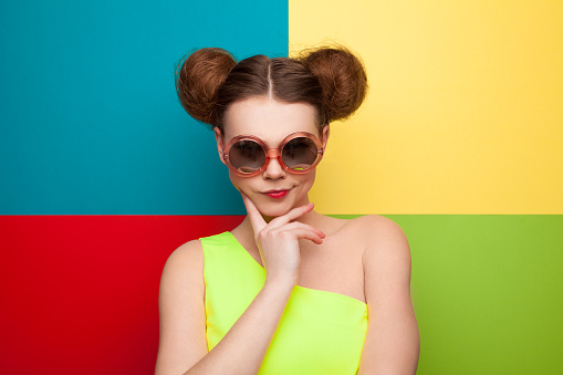 Young girl in summer outfit and sunglasses posing at camera on colorful background.