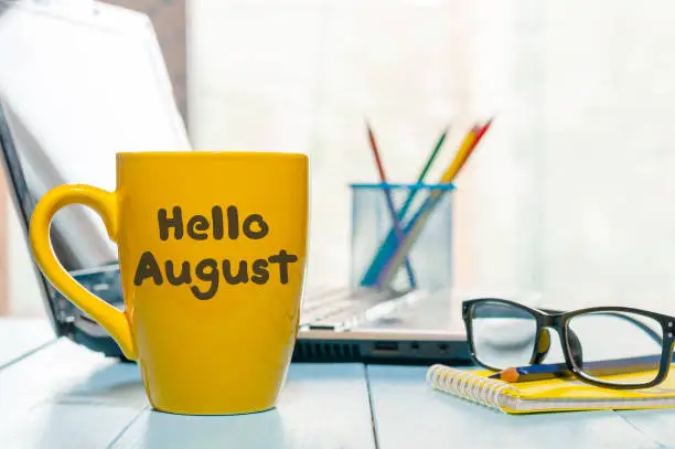 Photo of Hello August - inscription at yellow morning coffee or tea cup at business office background. Summer month, Calendar concept