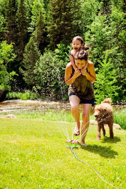 Millennial mother enjoying springkling water with baby daughter. Summer time is the perfect time to initiate little ones to sprinkling water games. This millennial mother, toddler and dog playing together running over it in the backyard nature. Vertical full length outdoors shot with copy space. This was taken in Quebec, Canada. dog swimsuit stock pictures, royalty-free photos & images
