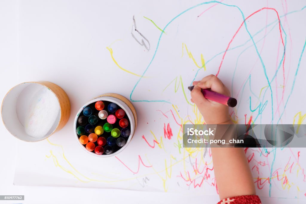 A hand of baby drawing lines and shapes with colorful crayons. Baby hold black color in her hand.Colorful crayons are in the box and put on the white paper. Crayon Stock Photo