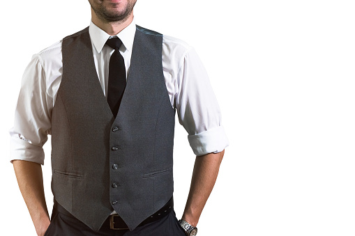 A young businessman in a white shirt with rolled up sleeves stands and keeps his hands in the pockets of his trousers. Hopster guy in a stylish gray waistcoat