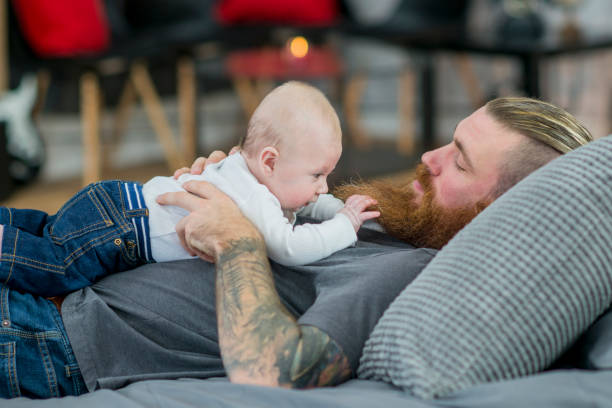 Napping With Baby An alternative-style father is lying down with his baby on top of him. The father has piercings, tattoos and a long beard. The baby is wearing regular clothing. chest tattoos for men designs stock pictures, royalty-free photos & images