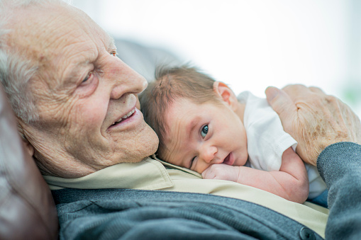 A Caucasian grandfather is indoors with his newborn grandson. They are both wearing casual clothing.  The grandfather is lying down with the baby on top of him.