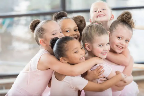 A group of young smiling ballerinas hug each other in a large huddle.  Three of them are looking at the camera.