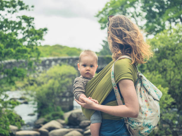 A young mother with a baby in a carrier sling is standing by a bridge in nature
