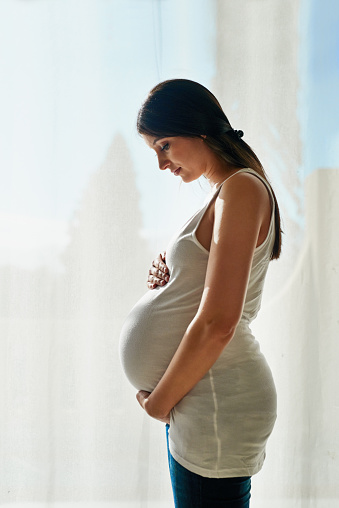 Shot of a pregnant woman cradling her belly while standing in front of a window at home