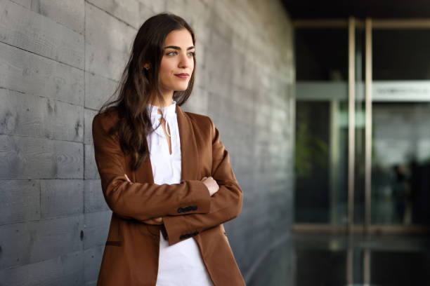 Young businesswoman standing outside of office building. Young businesswoman standing outside of office building. Beautiful woman wearing formal wear. Young girl with brown jacket and trousers. Blazer stock pictures, royalty-free photos & images