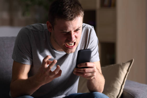 Angry man with a mobile phone at home Single angry person with a mobile phone sitting on a sofa in the living room in a house indoor with a dark background furious stock pictures, royalty-free photos & images