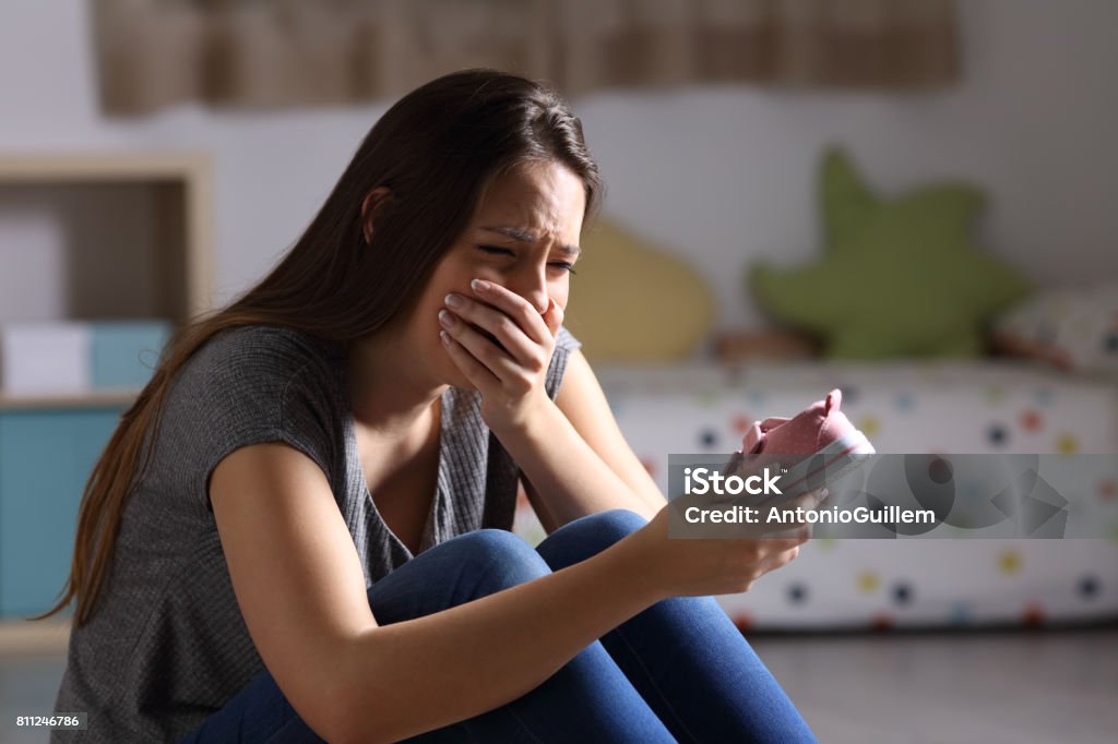 Sad mother missing her daughter Sad mother missing her daughter holding a little shoe sitting on the floor of the bedroom in a house interior with a dark background Grief Stock Photo