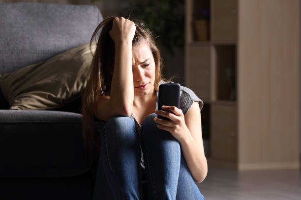 Sad teen checking phone at home Front view of a sad teen checking phone sitting on the floor in the living room at home with a dark background online bullying stock pictures, royalty-free photos & images