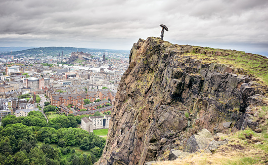 A woman standing above Salisbury Crags, in Holyrood Park, holding an umbrella for shelter as she looks over the city of Edinburgh.