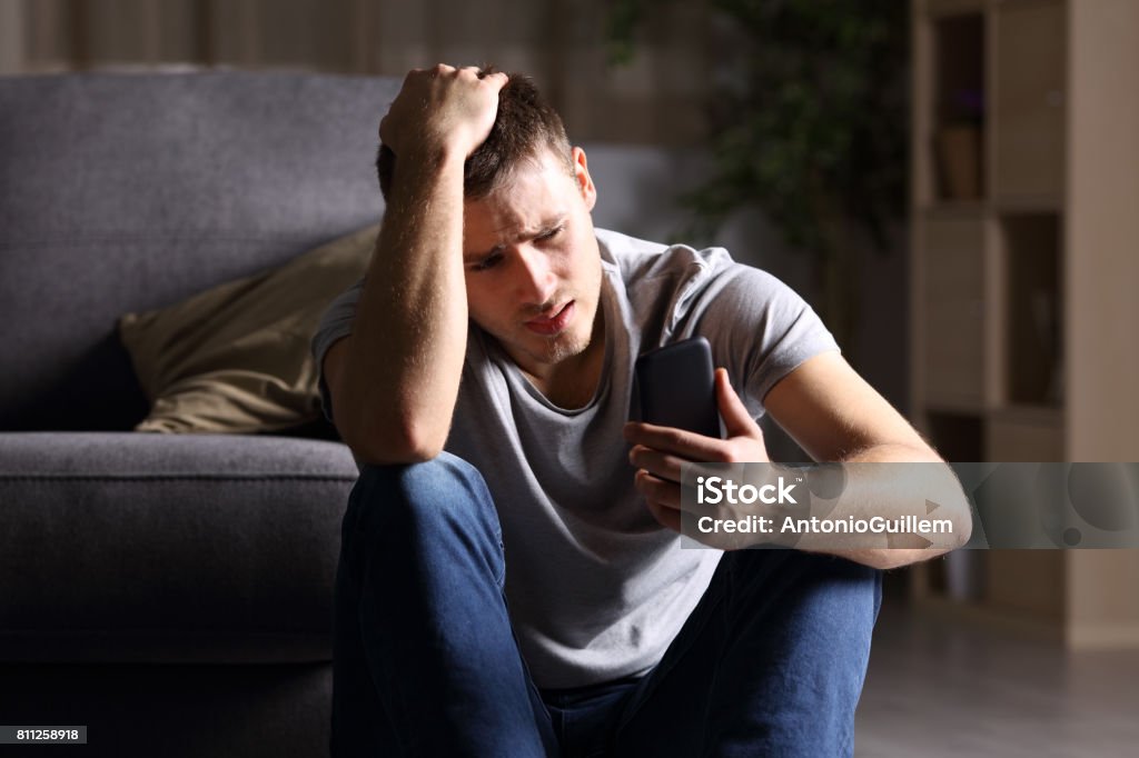 Sad man checking mobile phone Single sad man checking mobile phone sitting on the floor in the living room at home with a dark background Men Stock Photo