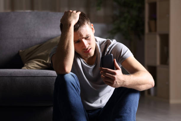 Sad man checking mobile phone Single sad man checking mobile phone sitting on the floor in the living room at home with a dark background relationship breakup photos stock pictures, royalty-free photos & images