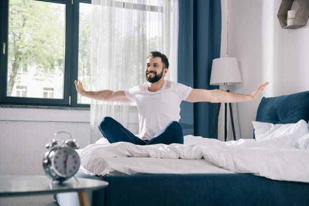 istock Smiling young man in pajamas stretching while sitting on bed at morning 811259328