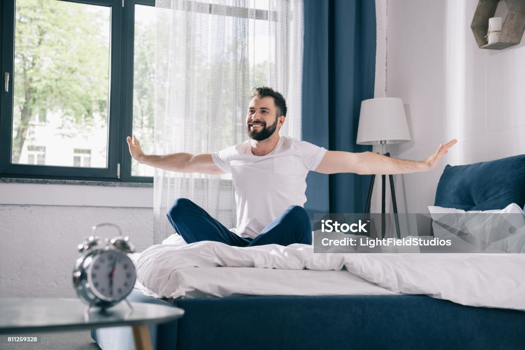 Smiling young man in pajamas stretching while sitting on bed at morning Waking up Stock Photo