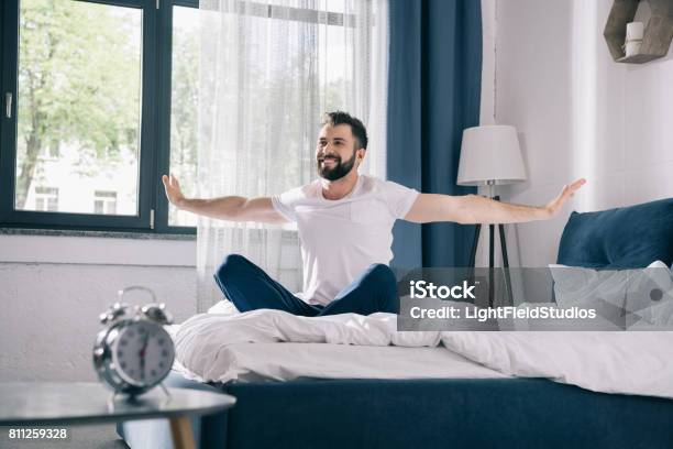 Smiling Young Man In Pajamas Stretching While Sitting On Bed At Morning Stock Photo - Download Image Now