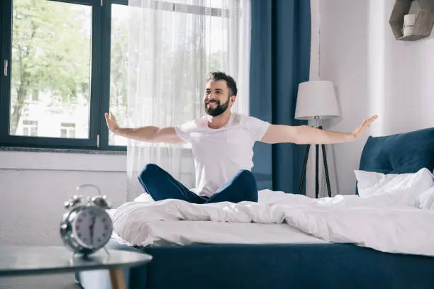 Photo of Smiling young man in pajamas stretching while sitting on bed at morning