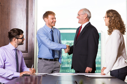 Cheerful business partners shaking hands after meeting, their assistant looking at them. Smiling senior boss congratulating colleague in board room. Results of conference concept