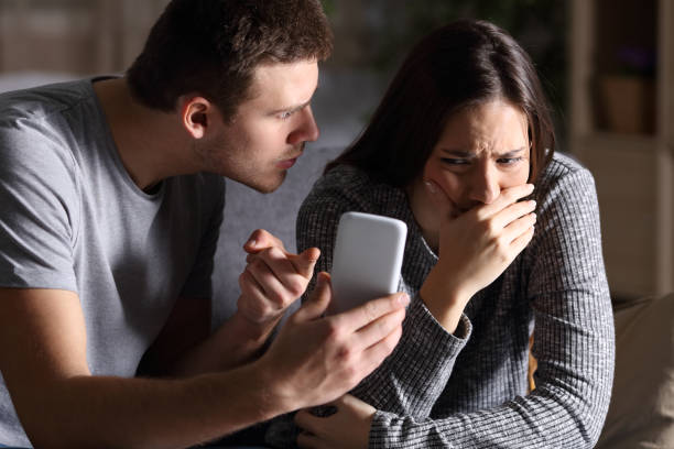 Boyfriend show phone to his cheater girlfriend Boyfriend asking for an explanation to his cheater sad girlfriend sitting on a couch in the living room in a house interior with a dark background suspicious message stock pictures, royalty-free photos & images