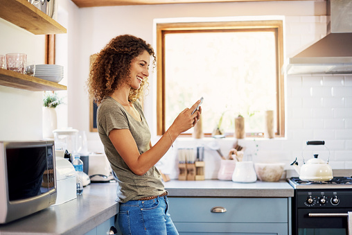 Shot of a happy young woman using her smartphone while relaxing in her kitchen at home