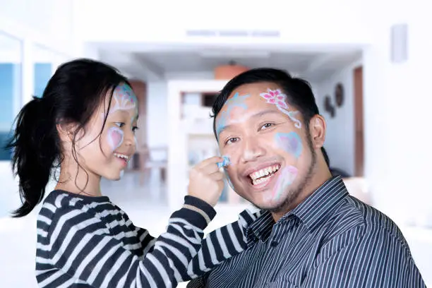 Picture of young father and his daughter playing with a crayon while drawing their face at home