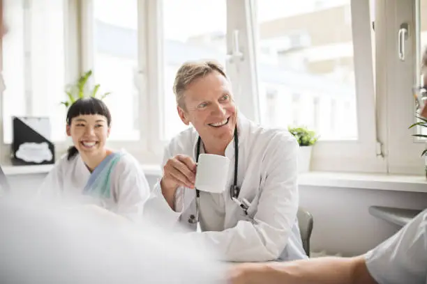 Happy doctor talking with his team during coffee break at hospital. Medics enjoying coffee during work break.