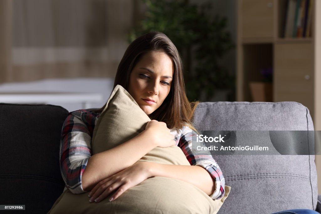 Sad girl embracing a pillow at home Front view of a sad single girl embracing a pillow sitting on a couch in the living room at home with a dark light in the background Depression - Sadness Stock Photo