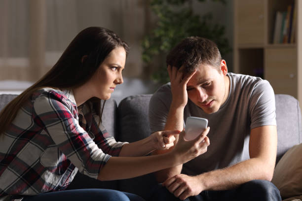 Girl asking for explanation of her cheater boyfriend Girlfriend asking for explanation of her boyfriend sitting on a couch in the living room in a house interior with a dark background suspicious message stock pictures, royalty-free photos & images