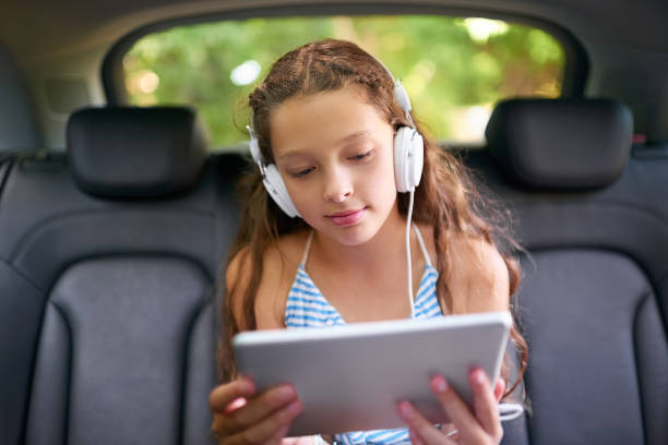 Keeping the car trip an entertained one Shot of a young girl sitting in a car wearing headphones and using a digital tablet entertainment equipment stock pictures, royalty-free photos & images