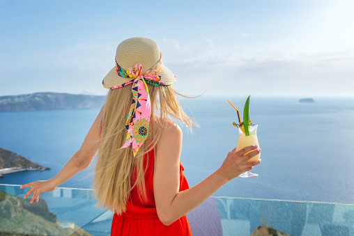 Horizontal color rear view image of elegant woman holding cocktail - pina colada and looking at sea view on balcony.