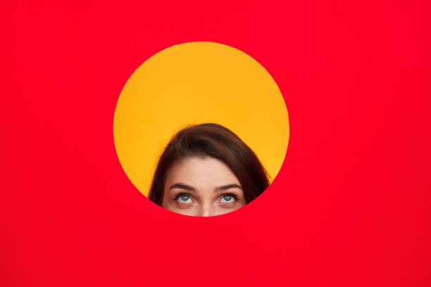 Young woman standing behind paper with cut circle looking out.