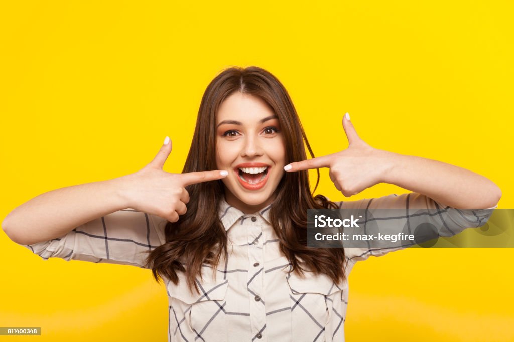 Woman smiling and pointing at mouth Cheerful girl pointing on her opened mouth and looking at camera on the yellow background. Smiling Stock Photo