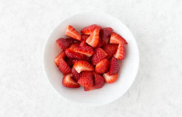 Closeup of cut strawberries in a white porcelain bowl isolated on white marble background with lots of copy space.