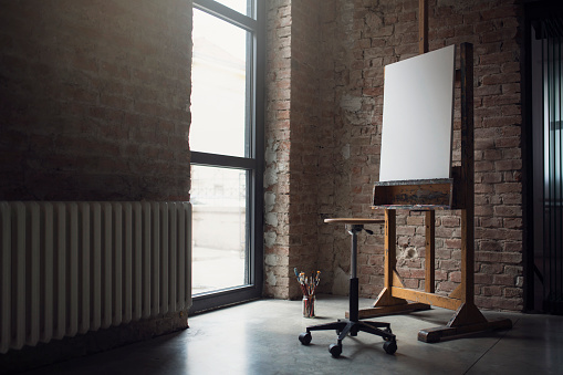 White canvas on painting easel, stool and paintbrushes in an artistic studio.