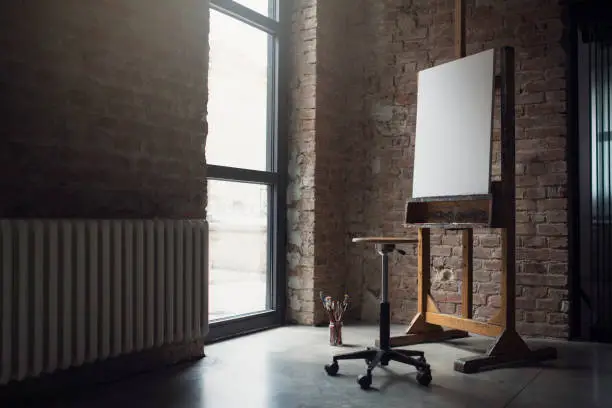 White canvas on painting easel, stool and paintbrushes in an artistic studio.