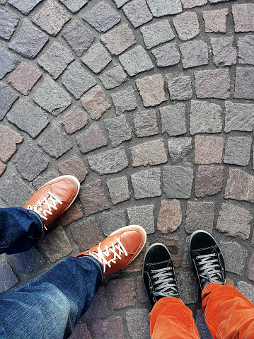 Legs and feet of two people wearing sneakers standing on cobblestone pavement from above with copy space