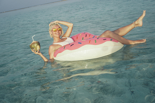 Young, attractive and beautiful woman is taking a selfie while floating in float in the ocean. Float is pink and yellow and have shape of doughnut witch is missing one part, something like one bite. She is wearing white swimsuit and orange sunglasses. In her left hand she is holding her hair and in right she have coconut fruit with straw. One leg is in the air and other one is in the water. Smile on her face is sign that she is relaxing and enjoying in this quite environment. Her short blonde hair is wet. Water is crystal clear and turquoise colored and sky is blue without clouds.