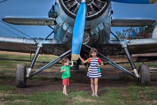 Two kids: preschool and elementary age are posing on a meadow. Agricultural biplane is behind them. Kids are holding huge propeller of the plane.
