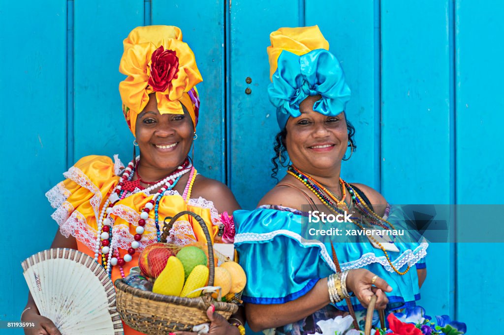 Portrait of women in Cuban traditional dresses Portrait of happy Cuban women standing against blue wooden wall. Smiling mature women are in traditional dresses. They are with fruit basket and hand fan. Cuba Stock Photo