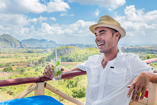 Cheerful mid adult man holding mojito glass against Valle De Vinales. Cuban male is enjoying vacation at outdoor restaurant in Cuba. He is wearing casuals.