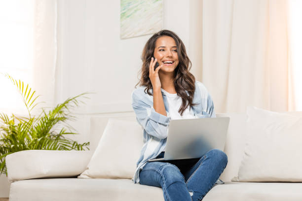 istock Smiling young woman talking on smartphone and using laptop while sitting on sofa at home 811970614