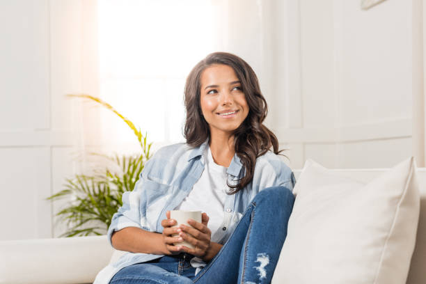 istock Pensive smiling young woman holding cup while sitting on sofa and looking away at home 811987652