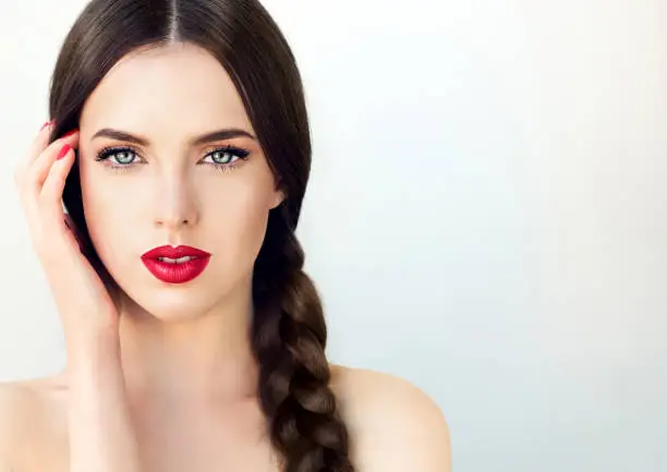 Close up portrait of beautiful  young brunette woman-model  with  long braided hair. Pigtail hairstyle. Bright blue eyes, red lipstick on the lips and red  manicure on the nails.