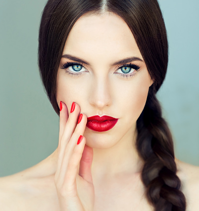 Close up portrait of beautiful  young brunette woman-model  with  long braided hair. Pigtail hairstyle. Bright blue eyes, red lipstick on the lips and red  manicure on the nails.