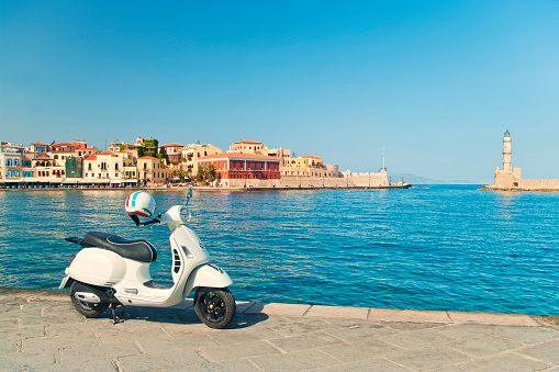 cross-processed image of white retro style scooter parked in harbour in Chania on sunny summer day with old city and venetian light-house at background, Crete